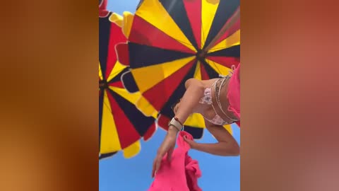 Media: Video of a woman with light skin, wearing a pink dress, holding a vibrant umbrella with alternating red, yellow, and black stripes against a bright blue sky.