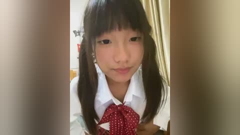 Media: A video of an Asian girl with straight black hair in pigtails, wearing a white shirt with a red polka-dot bow tie. Background shows a beige curtain and blurred indoor setting.