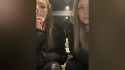 Media: Video of two women with long brown hair, wearing black coats, sitting close together, looking at each other, indoors with dim lighting and blurred background.