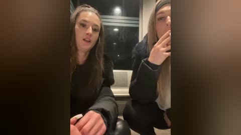 Media: Video of two young women, one with long hair, wearing black, sitting inside a darkened train. The woman on the left has a concerned expression while the other covers her mouth with a finger.