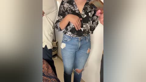 Media: Video of a woman with light brown skin, wearing a black floral blouse and light blue distressed jeans, taking a selfie in a mirror.