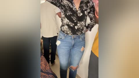 Media: Video of a person wearing a black floral blouse and distressed light blue jeans with white patches, standing in a brightly lit, modern indoor setting.