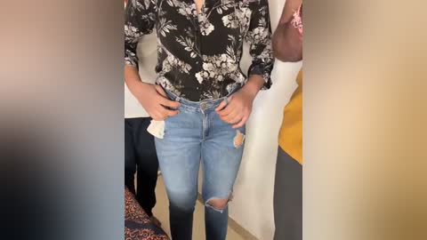 Media: Video of a person in light blue ripped jeans and a black floral blouse, standing in a hallway.