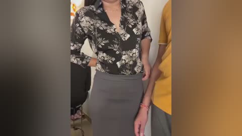 Media: A video shows a woman in a black floral blouse and high-waisted gray pencil skirt, standing in a room with a yellow-shirted man partially visible.