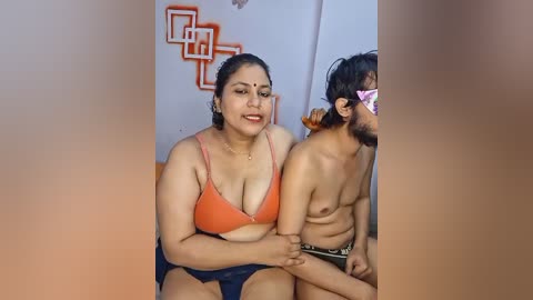 Media: Video of a plus-sized woman with dark skin and a large breast, wearing an orange bra and shorts, seated next to a shirtless, skinny man with glasses and a beard.