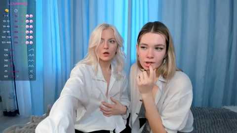 Media: Video of two young women with fair skin, one with blonde hair, the other with straight, shoulder-length brown hair, both wearing white shirts. They sit on a grey couch in front of blue curtains.