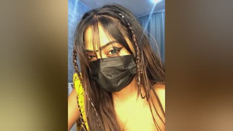 Media: Video of a young woman with long, dark brown hair in braids, wearing a black face mask, yellow strap, and a sleeveless top, indoors with a blurred background.