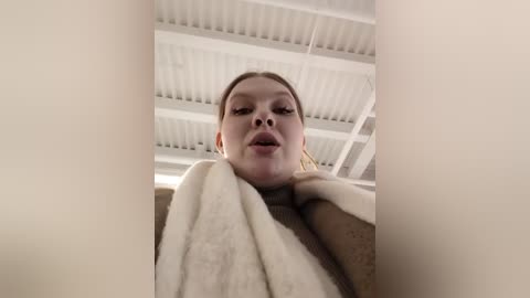 Media: Video of a Caucasian woman with light skin, brown hair, and a neutral expression, wearing a beige coat, standing in a modern, white-tiled room with a high ceiling.