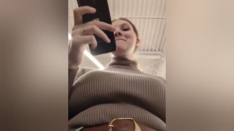 Media: Video of a young woman with light skin, brown hair, and large breasts, wearing a ribbed brown sweater and a brown belt, taking a selfie in a modern, industrial room with white walls and corrugated metal ceiling.