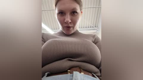 Media: Video of a light-skinned woman with blonde hair, wearing a tight, ribbed, beige turtleneck sweater and high-waisted light blue jeans, standing in an industrial warehouse with corrugated metal walls and fluorescent lighting.