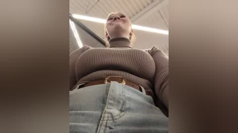 Media: A video of a young woman in a brown ribbed turtleneck sweater and high-waisted light blue jeans, taken from a low angle, emphasizing her large breasts and curvy hips.