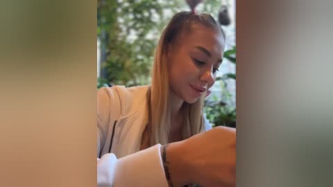 Media: A video of a young woman with light skin and blonde hair in a ponytail, wearing a white jacket, seated at a table, smiling at someone off-frame, surrounded by blurred greenery.