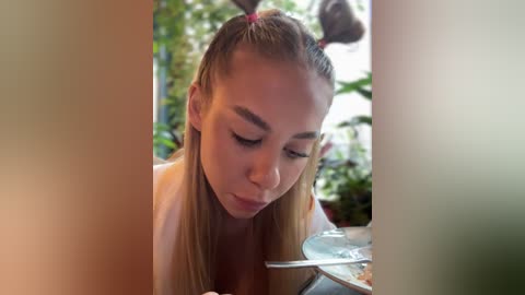 Media: Video of a young Asian woman with straight, blonde hair tied in two high pigtails, wearing a white shirt, intently reading a book, in a blurred outdoor setting with greenery.