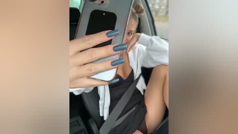 Media: Video of a woman with fair skin and blue nails, wearing a white lab coat and black top, taking a selfie in a car, seatbelt visible.