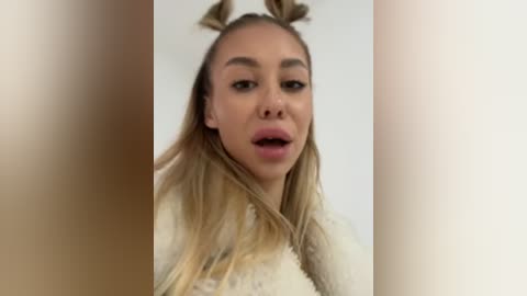 Media: Video of a young woman with light skin, long blonde hair in two braids, wearing a cream sweater. She has a neutral expression and is in a well-lit indoor setting.