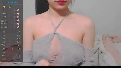 Media: A video of an Asian woman with fair skin, black hair, and red lipstick, wearing a sheer, off-shoulder gray dress, against a plain background.