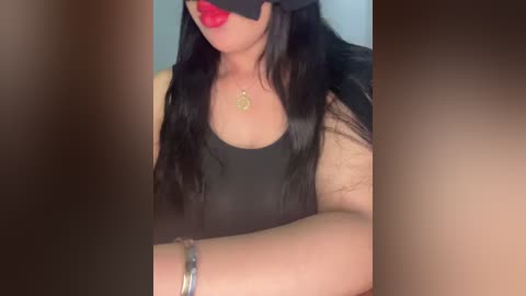 Media: Video of a woman with long black hair, wearing a black mask and sleeveless black top, red lipstick, and gold necklace, against a blurred background.