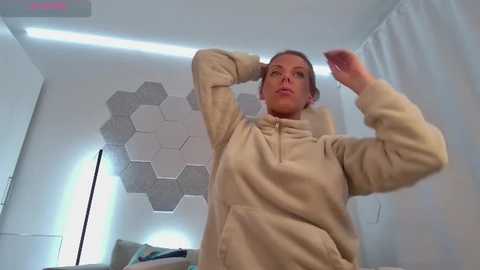 Media: Video of a woman in a beige hoodie, adjusting her hair, in a modern room with white walls, hexagonal light panel, and a tall floor lamp.