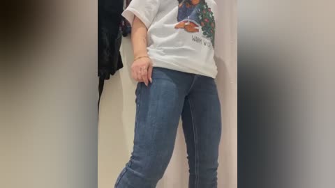 Media: Video of a woman in a white T-shirt with a graphic design and blue jeans, standing in a dressing room with a blurred background.