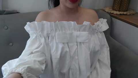 Media: A video of a woman with light skin, wearing a white, off-shoulder blouse with ruffled details, seated on a gray sofa. She has dark hair and red lipstick. Background includes a wooden table with a pineapple decor piece.