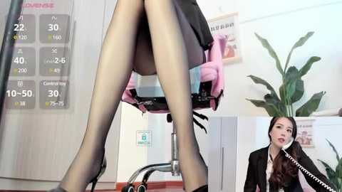 Media: Video of a woman in a black skirt, black stockings, and high heels sitting on a pink office chair, talking on a phone, with a desk calendar and potted plant in the background.