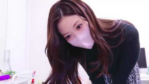 Media: Video of a young Asian woman with long brown hair, wearing a black sweater, white face mask, and floral skirt, bending over a desk in a bright, minimalist room.