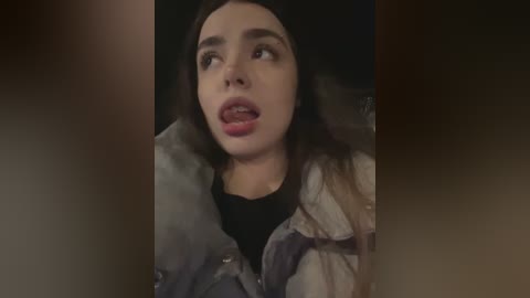 Media: Video of a young woman with fair skin and long dark hair, wearing a grey denim jacket over a black shirt, and a black beanie, with a surprised, open-mouthed expression.