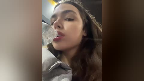 Media: Video of a young woman with long brown hair, light skin, and full lips, drinking from a clear plastic bottle in a dimly lit room.