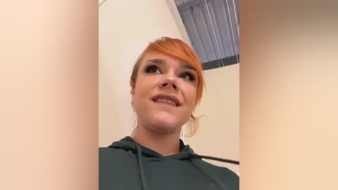 Media: Video of a smiling woman with light skin and orange hair, wearing dark makeup, standing in a beige room with a slanted ceiling and vertical blinds.