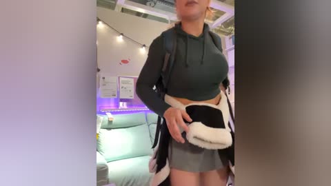 Media: Video of a woman wearing a black hoodie and grey skirt, carrying a white and black furry handbag, in a cozy, dimly-lit room with a teal couch, string lights, and framed artwork on the wall.