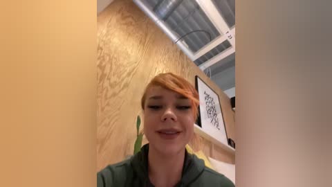 Media: A video of a young woman with light skin and red hair, smiling, standing in a modern, brightly lit room with wooden walls and exposed ceiling beams.