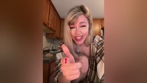 Media: Video of a smiling, fair-skinned blonde woman in a plaid shirt giving a thumbs-up in a cozy kitchen with wooden cabinets and beige countertops.