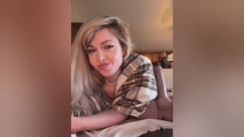 Media: Video of a young woman with long, straight blonde hair, wearing a plaid shirt, lying on a bed, smiling softly. The background features a wooden dresser and warm lighting.