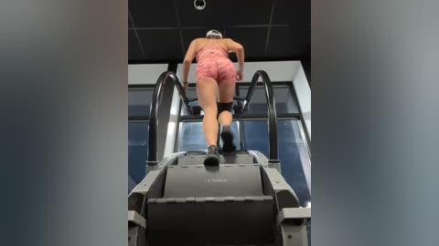 Media: A video of a fit, fair-skinned woman in pink lace shorts and a sports bra ascending an escalator in a modern, well-lit building. She has a headband and black sneakers, with a clear focus on her athletic physique.