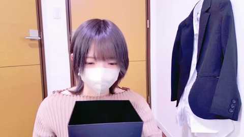 Media: Video of an Asian woman with straight, shoulder-length black hair wearing a beige sweater, face mask, and looking at a black tablet in a modern, minimalist room with a wooden door and a dark suit hanging on a hook.