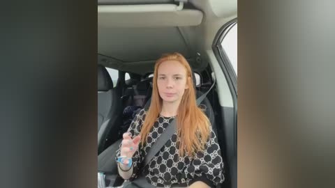 Media: Video of a young woman with long, straight red hair, wearing a black and white patterned top, driving a car with a seatbelt and water bottle in hand.