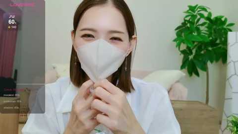 Media: Video of an Asian woman with shoulder-length brown hair, wearing a white surgical mask, holding a tissue. She's in a room with a potted plant and a bed in the background.