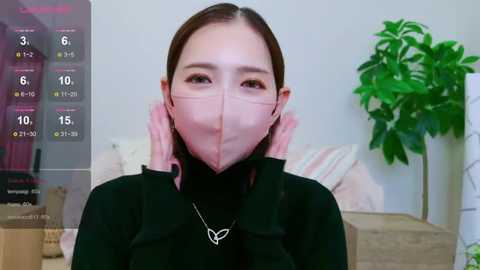 Media: Video of an East Asian woman with fair skin, brown hair, and dark eyes wearing a black mask, black top, and necklace, in a modern living room with a green plant.