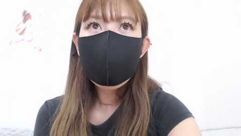 Media: Video of an East Asian woman with long brown hair, wearing a black face mask and black t-shirt, set against a plain white background.