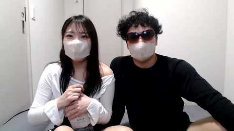 Media: Video of two masked Asian individuals, a woman with long black hair and a man with curly hair, both wearing white face masks and black clothes, sitting in a white-walled room.