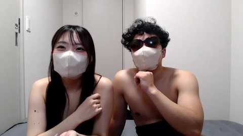 Media: Video of a slender Asian woman with long black hair and a mask, and a muscular Asian man with curly black hair and dark sunglasses, both shirtless, in a dimly lit room with white walls.