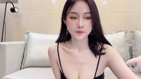 Media: Video of an East Asian woman with long black hair, fair skin, and subtle makeup, wearing a black spaghetti strap top, sitting on a beige couch with light pillows.