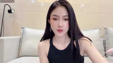 Media: Video of an East Asian woman with fair skin, long black hair, and light makeup, wearing a black sleeveless top, sitting on a light-colored couch in a modern, minimalist room.