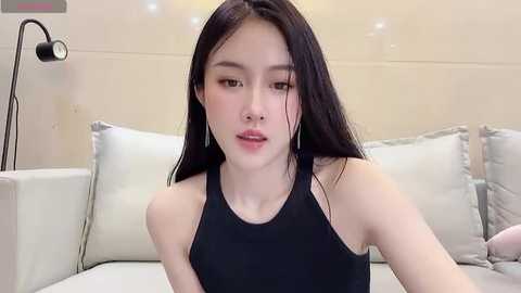 Media: Video of a young East Asian woman with long black hair, wearing a black tank top, sitting on a white couch with pillows. Background features a beige wall and a black desk lamp.