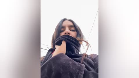 Media: Video of a young woman with medium brown skin, wearing a dark hooded jacket, partially covering her face. She has long, straight brown hair. Background is blurred with a white sky and power lines.