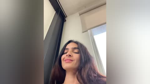 Media: A video of a young woman with long dark hair, fair skin, and closed eyes, smiling slightly, in a modern, light-colored room with a window and a beige curtain.