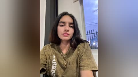 Media: Video of a young woman with long dark hair, wearing a beige t-shirt, sitting indoors with a window showing a cityscape in the background.