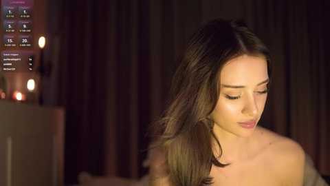 Media: Video of a serene, fair-skinned woman with long, straight brown hair, wearing a strapless top, sitting indoors by a dimly lit window with curtains.