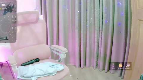 Media: Video of a modern bedroom with a plush pink chair, a blue blanket, and a black vibrator on the floor. Pastel curtains with star patterns, a TV screen, and a small table.