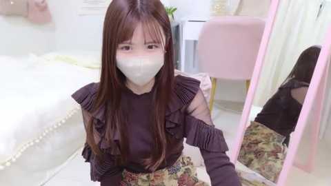 Media: Video of an Asian woman with long, straight brown hair, wearing a dark purple ruffled blouse and a white face mask, kneeling in a pink-walled bedroom with floral bedding and a white vanity table.